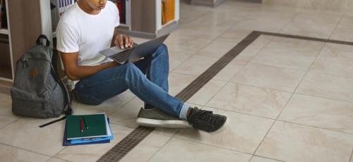Young afro guy holding laptop, sitting on floor at library